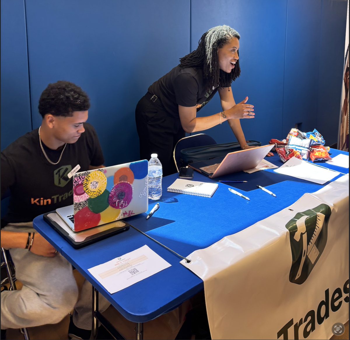 KinTrades staff at a job fair booth with a KinTrades banner, laptops, and sign-up sheets on the table.